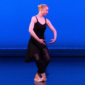 Susie Butler performs a plié in fifth position with arms in first position on the stage wearing a black leotard, skirt, and tights in 2023