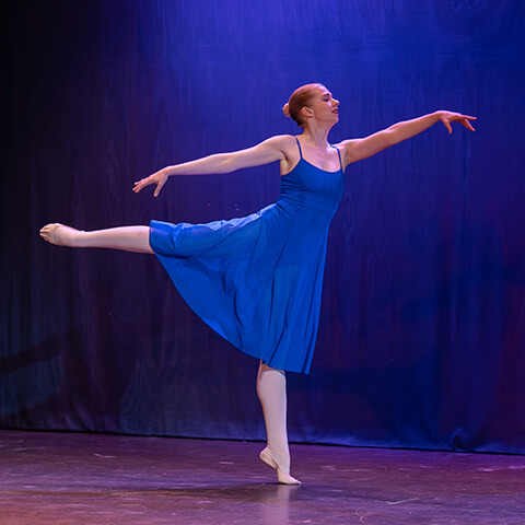 Susie performs an arabesque on stage in a flowy royal blue ballet dress